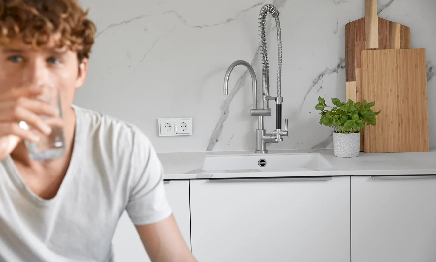 Ein Mann mit braunen Haaren und weißem T-Shirt trinkt ein Glas Wasser in einer weißen Küche.