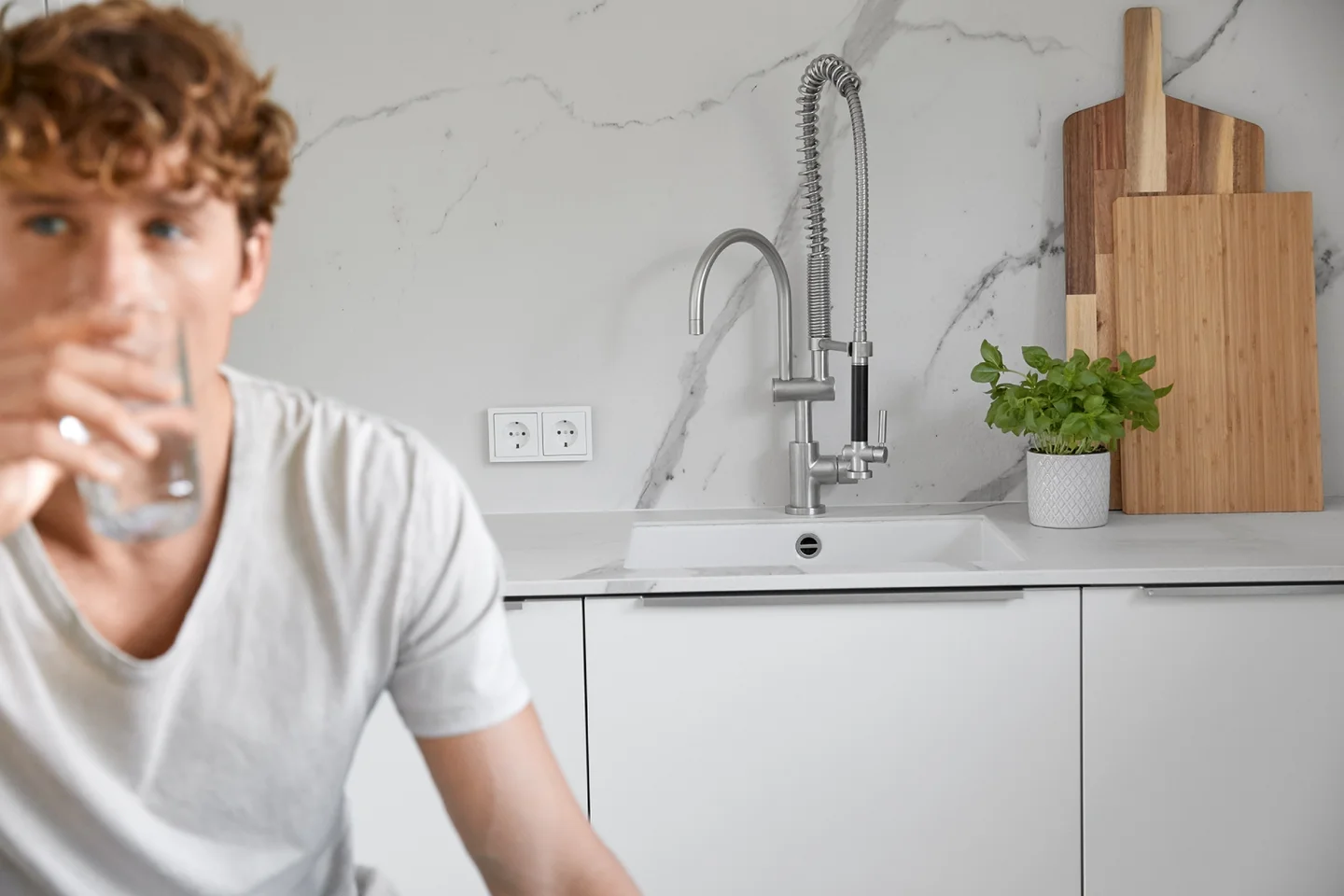 Ein Mann mit braunen Haaren und weißem T-Shirt trinkt ein Glas Wasser in einer weißen Küche.