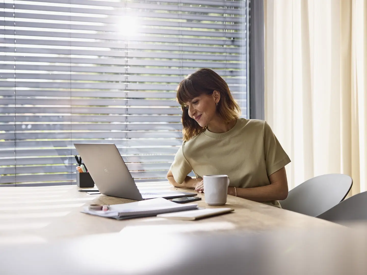 Frau sitzt an einem Tisch, schaut auf einen Laptop und hält eine Tasse. Notizblätter und ein Smartphone liegen auf dem Tisch.