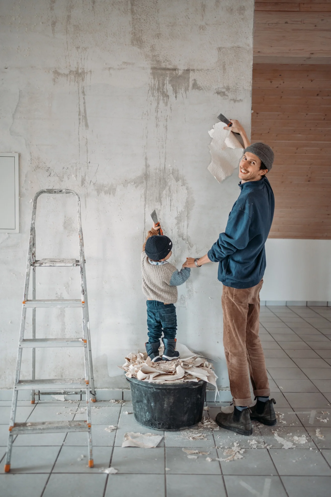Mann entfernt Tapete von einer Wand. Neben ihm steht ein kleines Kind erhöht auf einem Eimer, daneben eine Leiter.