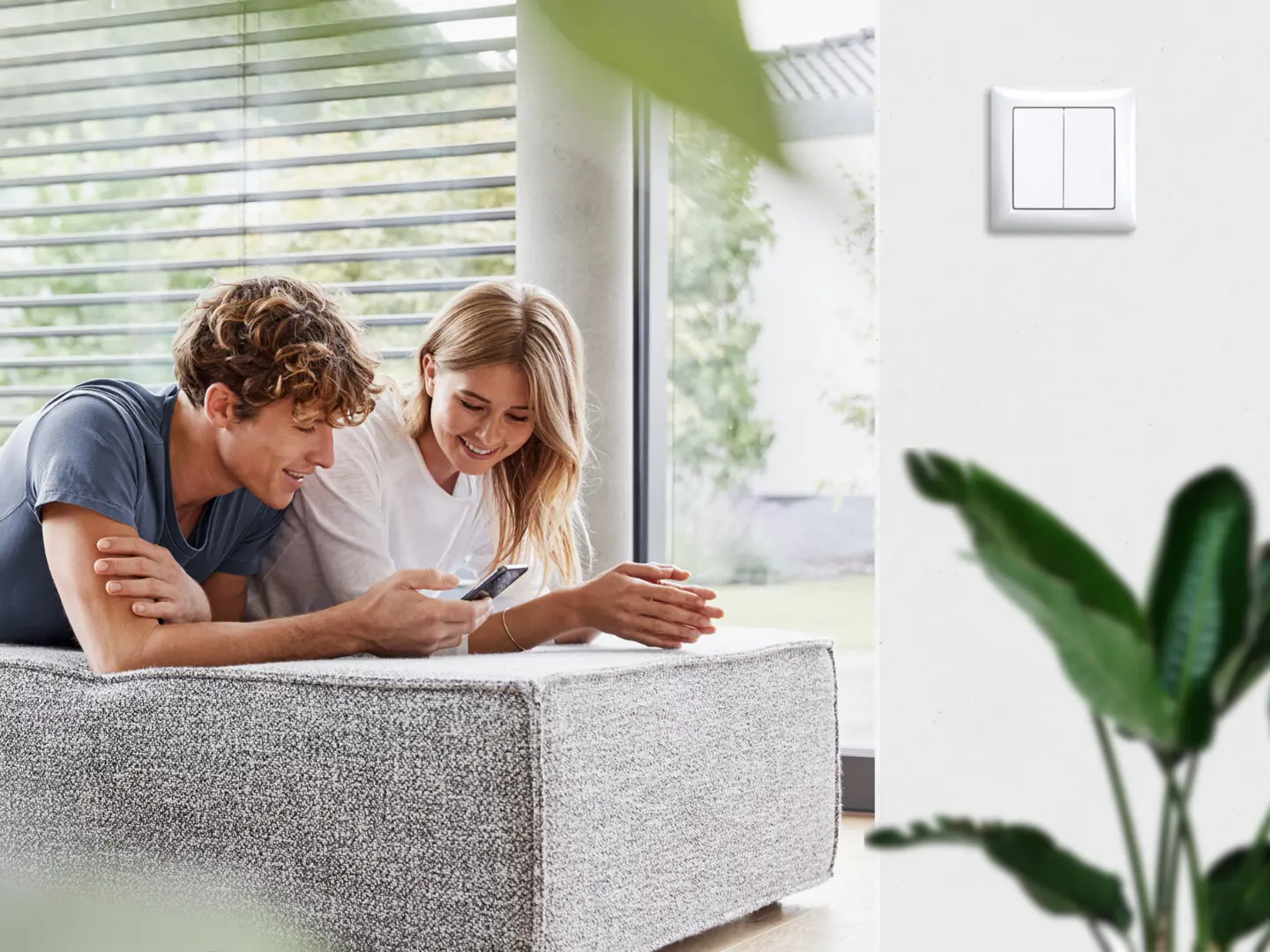 Ein Paar liegt entspannt auf einer Couch und bedient ein Tablet. An der Wand ist der Bluetooth Smart Switch sichtbar, der eine moderne Steuerungslösung bietet.