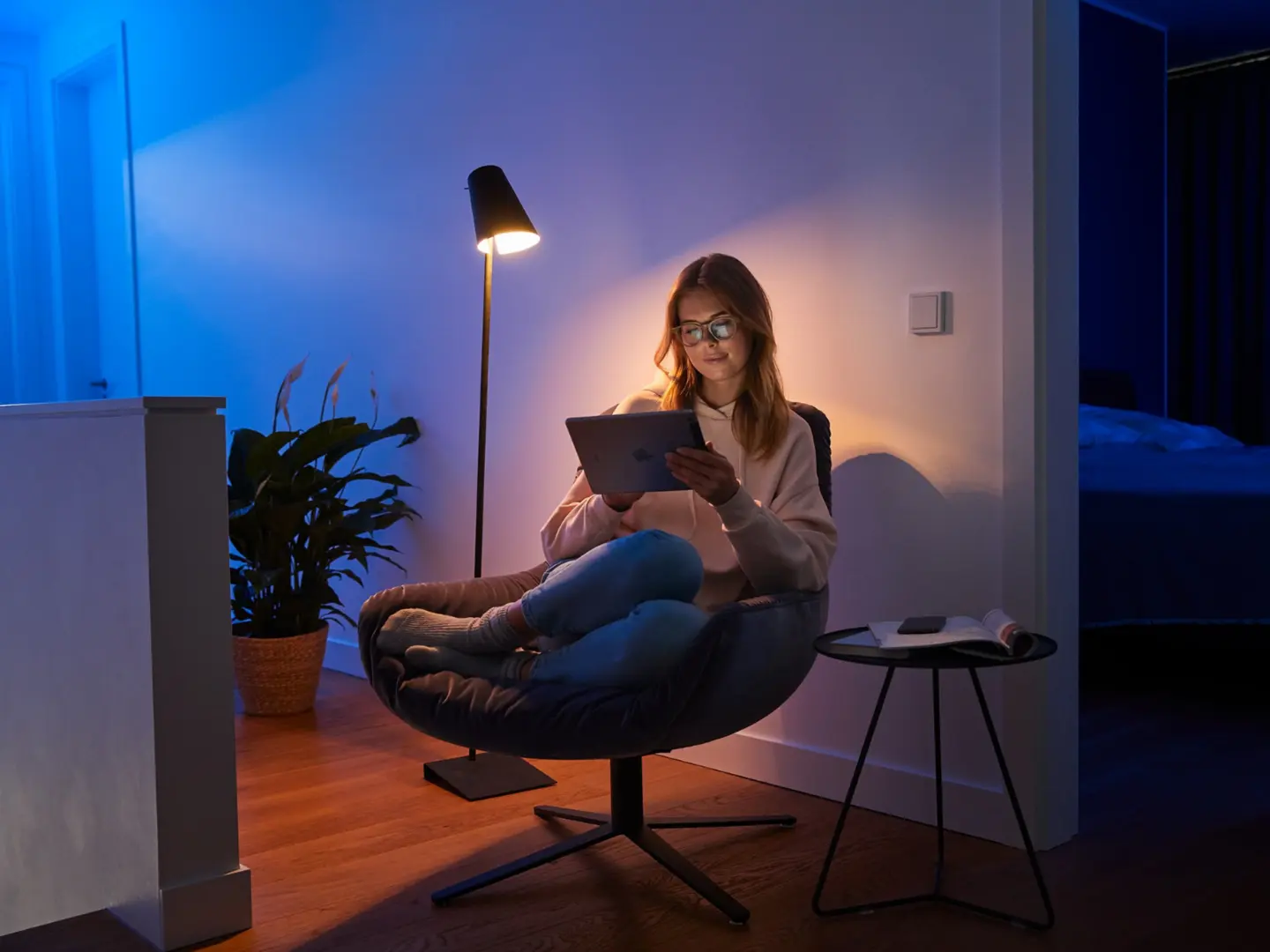 Eine Frau sitzt in einem Sessel und liest auf ihrem Tablet, während im Hintergrund die bunten Lichter von Philips Hue leuchten. Der Friends of Hue Schalter ist sichtbar und ergänzt das moderne Wohnambiente.