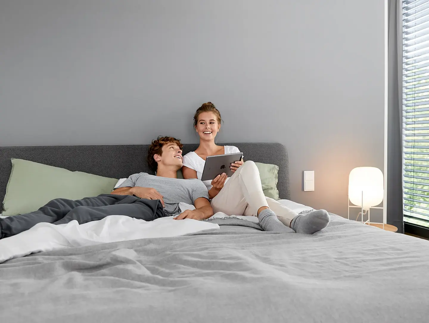 Ein Paar liegt im Bett und nutzt ein Tablet, um die Raumtemperatur in ihrem Smart Home zu steuern, was für Komfort und moderne Wohnlösungen steht.