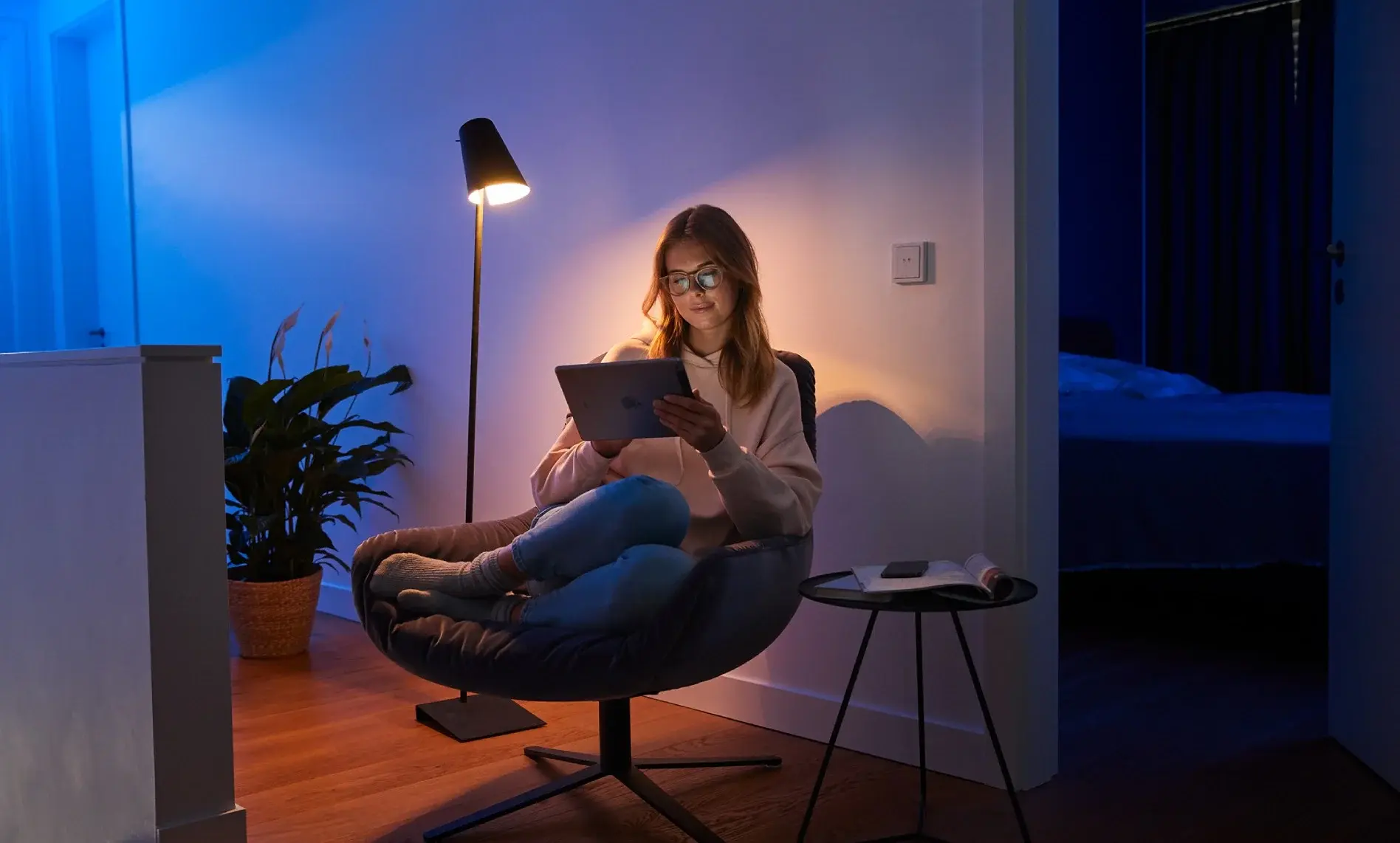 Eine Person mit Brille sitzt entspannt in einem Stuhl und liest auf einem Tablet, beleuchtet von einer Stehlampe.
