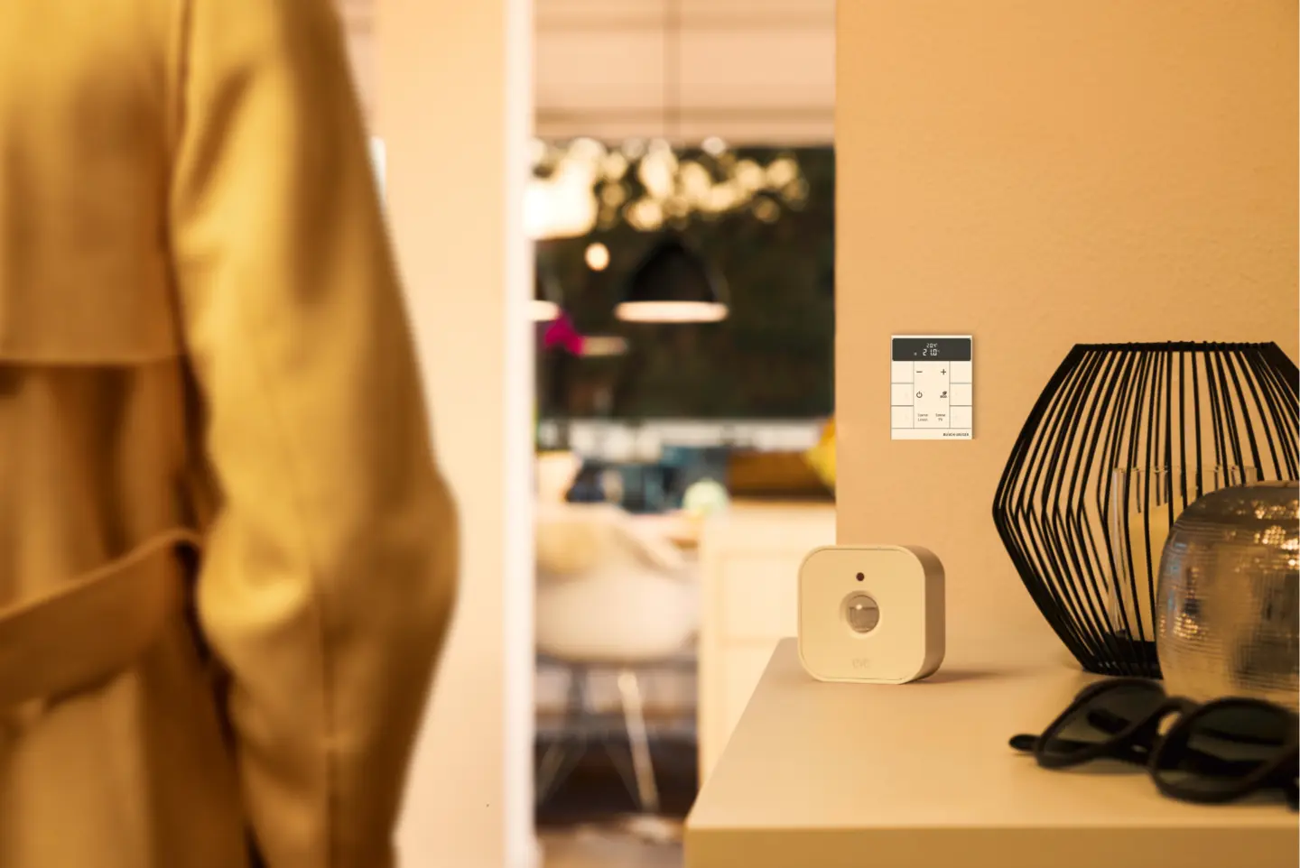Ein weißes Smart-Home-Gerät auf einem Tisch, neben einer modernen Lampe und einer Sonnenbrille, mit einer Person, die vorbeigeht.