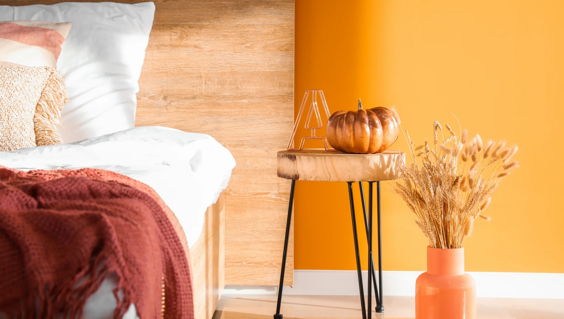 Ein Schlafzimmer mit einem Bett, einer Holzablage mit einem Kürbis und einer Vase mit Trockenblumen auf einem Boden mit orangener Wand.